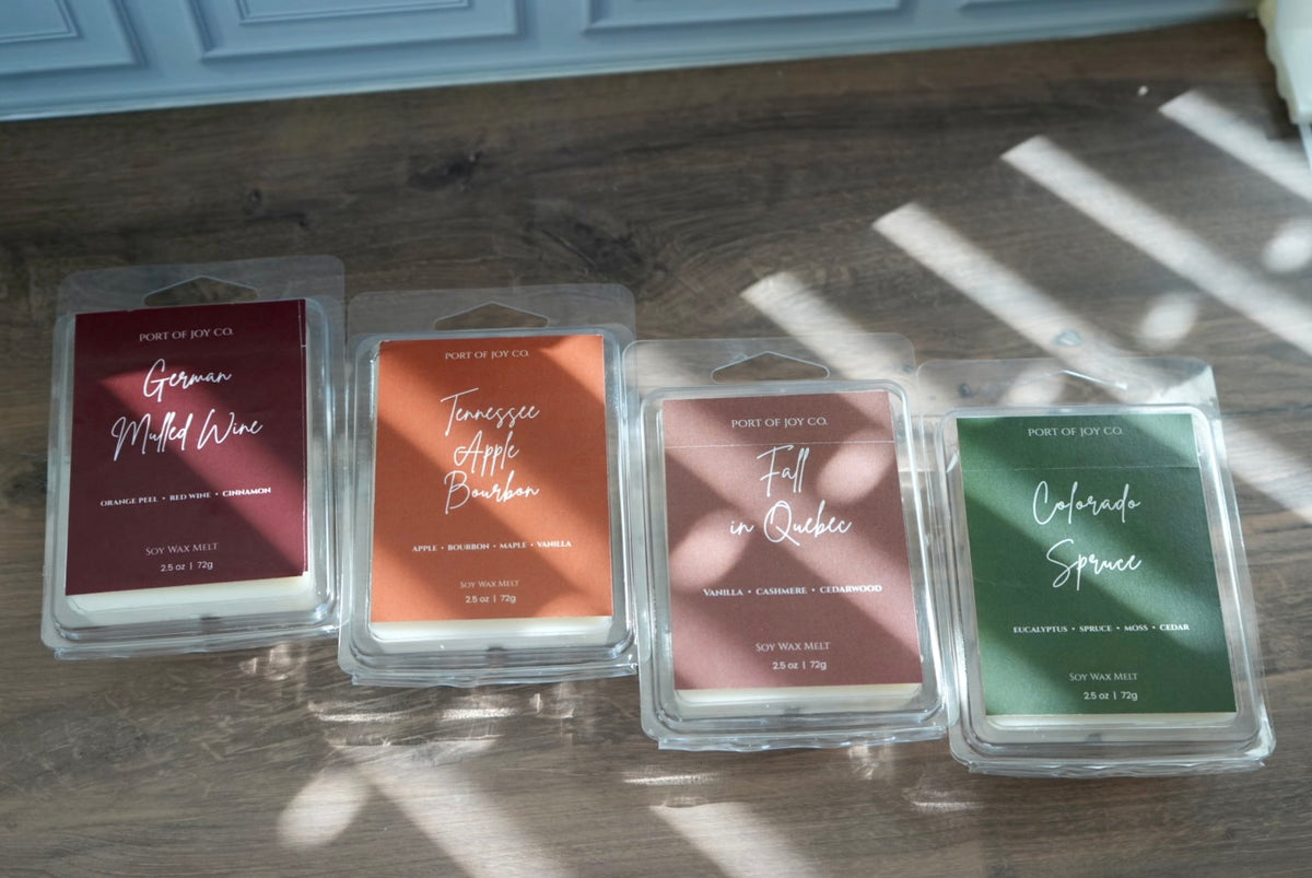Four wax melts in clear plastic clamshell packaging arranged on a wooden floor.