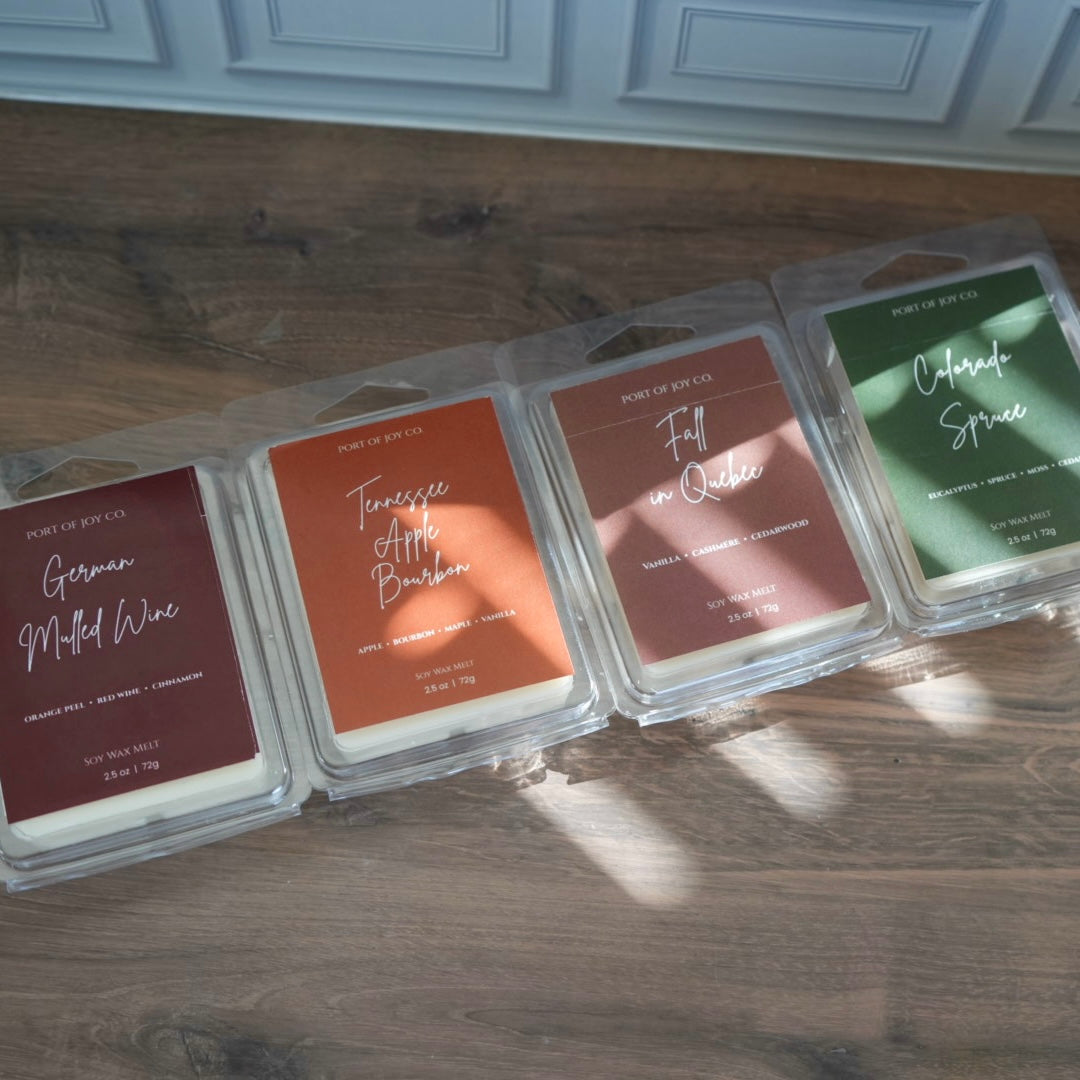 Four wax melts in clear plastic clamshell packages lined up on a wooden floor. fall collection 2026