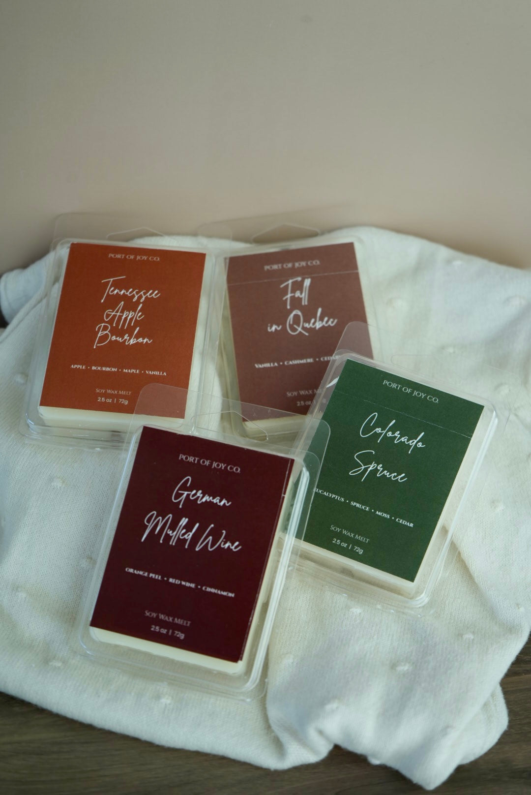 Four wax melts in clear plastic clamshell packaging arranged on a white textured fabric surface. 
