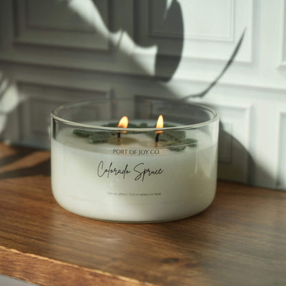 A lit double-wick candle in a clear glass jar sits on a wooden surface against a white paneled wall, with the label reading Port of Joy Co. and Colorado Spruce.