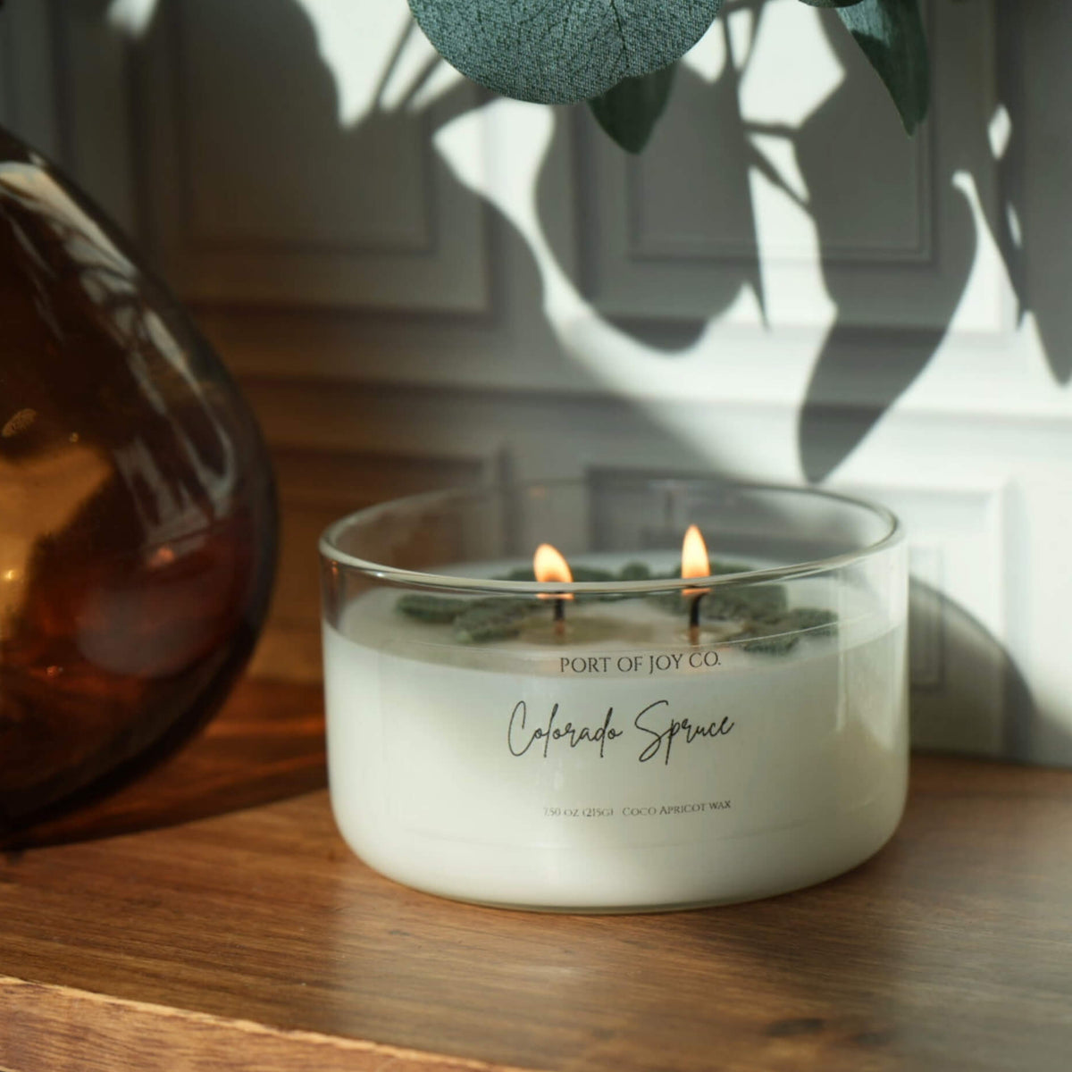 A double-wick scented candle in a clear glass jar sits on a wooden surface, lit with two flames. The label reads 'PORT OF JOY CO.