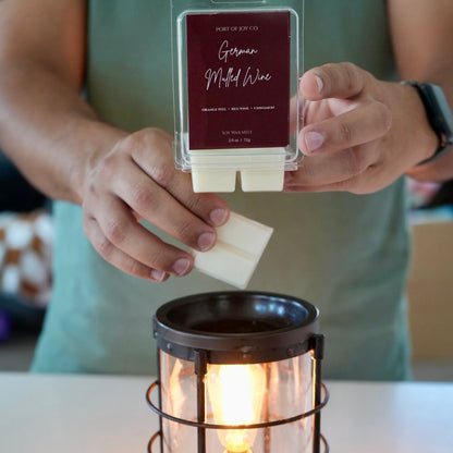 A person holds a clear wax melt package labeled 'Port of Joy Co. German Malted Wine' above a lit candle holder.