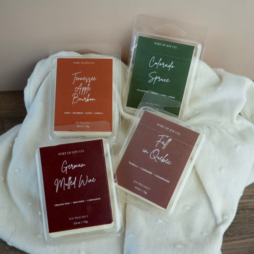 Four wax melts in clear clamshell packaging arranged on a white textured knit blanket. The labels read Port of Joy Co and the scents Tennessee Apple Bourbon, Colorado Spruce, German Milled Wine, and Fall in Quebec.