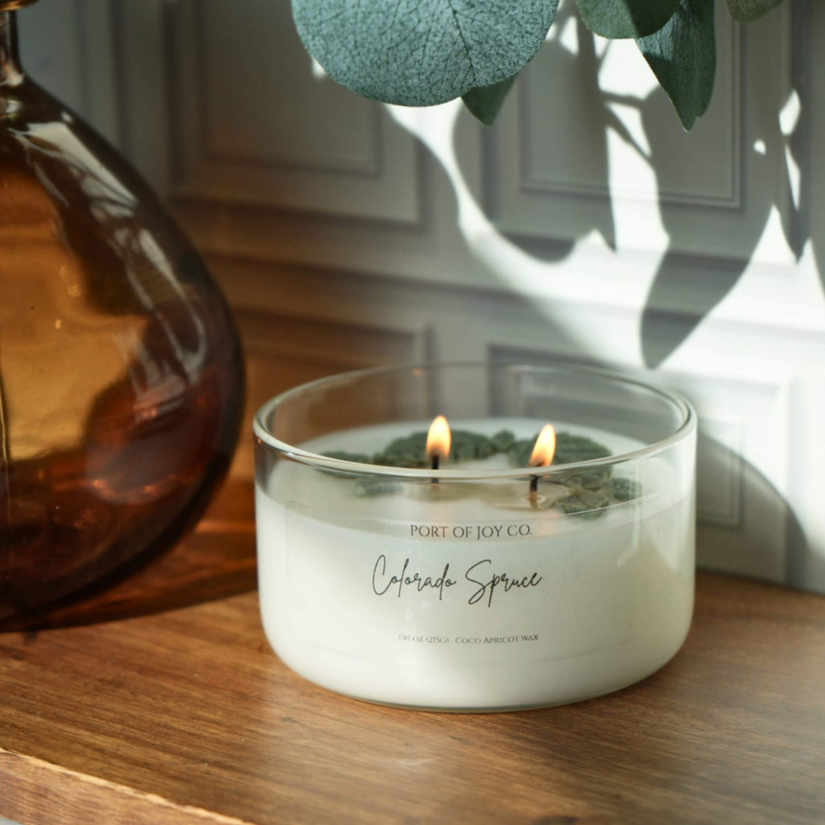 A lit double-wick scented candle in a clear glass jar sits on a wooden surface next to an amber glass bottle, with eucalyptus leaves casting shadows in the background. The label reads 'Port of Joy Co.