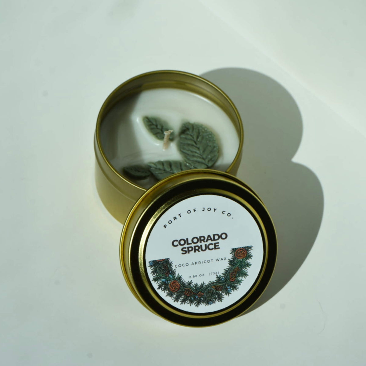 An open round metal candle tin with white wax and green leaf-shaped wicks, shown next to its lid. The lid label reads 'PORT OF JOY CO.