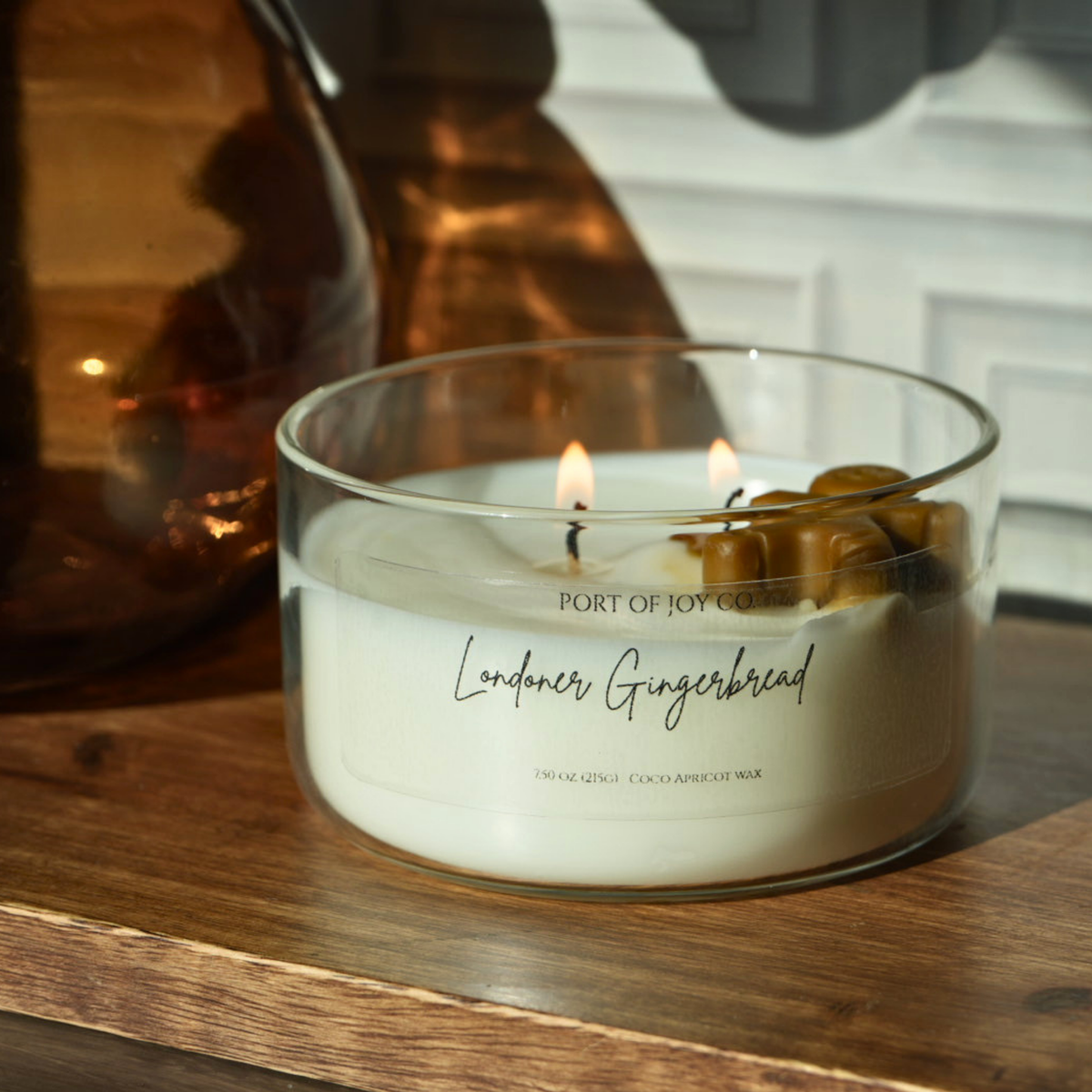 A lit scented candle in a clear glass jar sits on a wooden surface. The candle wax is white, with the label reading 'PORT OF JOY CANDLE Londoner Gingerbread.