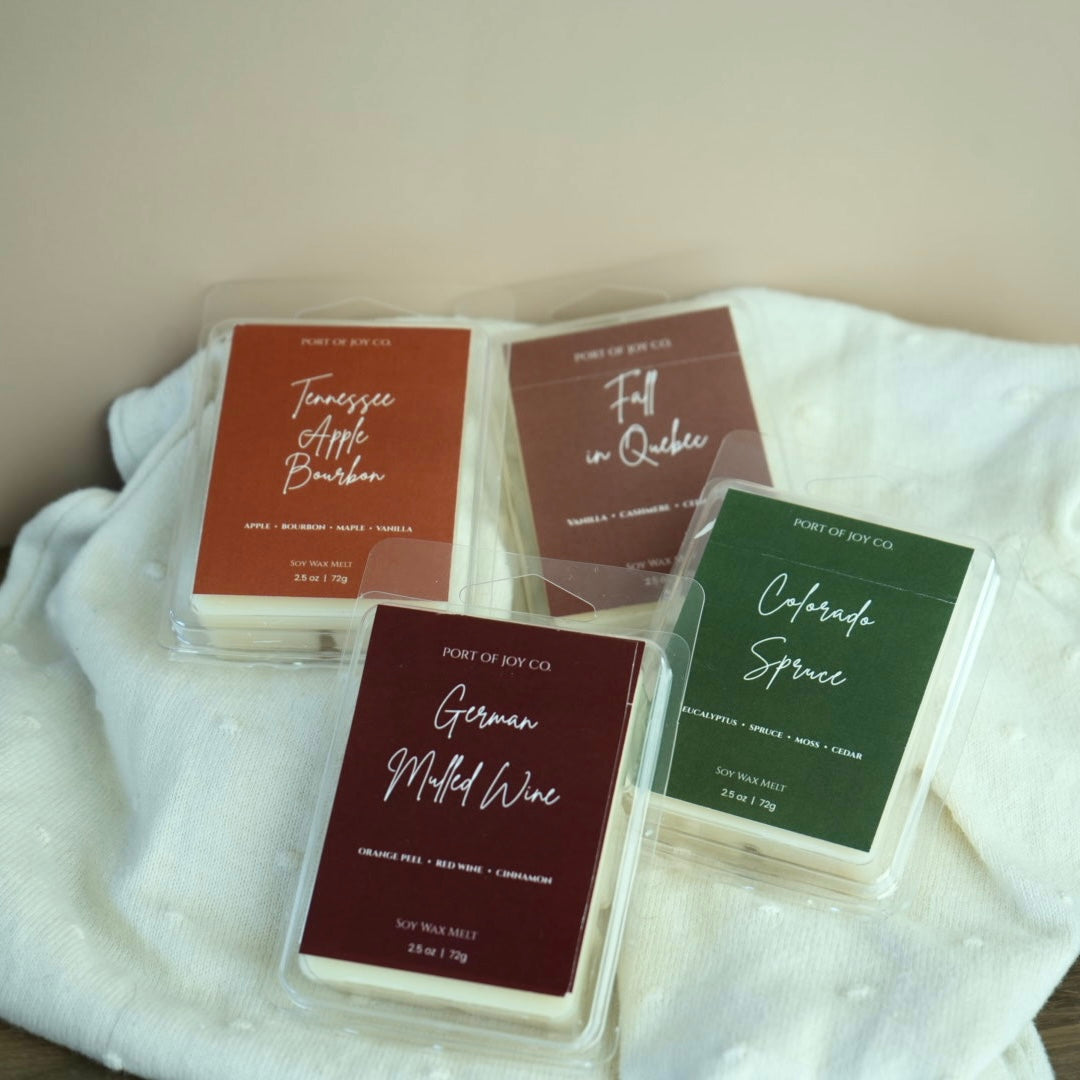 Four wax melts in clear plastic clamshell packaging arranged on a white textured fabric. 