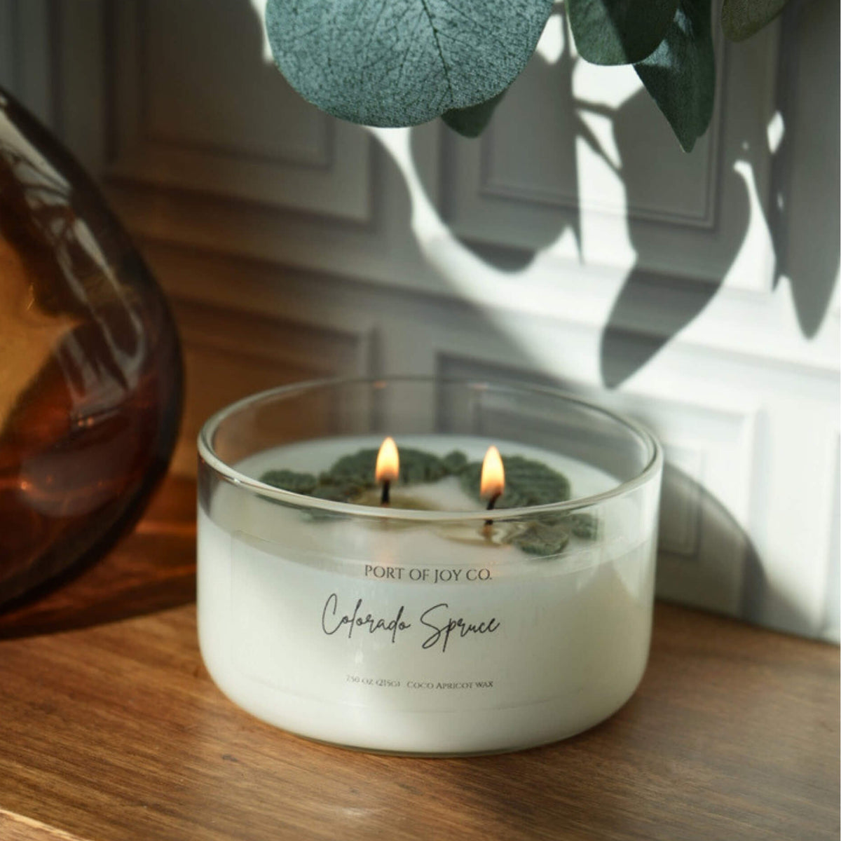 A double-wick candle in a clear glass jar labeled 'Port of Joy Co. Colorado Spruce' sits on a wooden surface with green leaves hanging overhead and casting shadows on a white paneled wall.