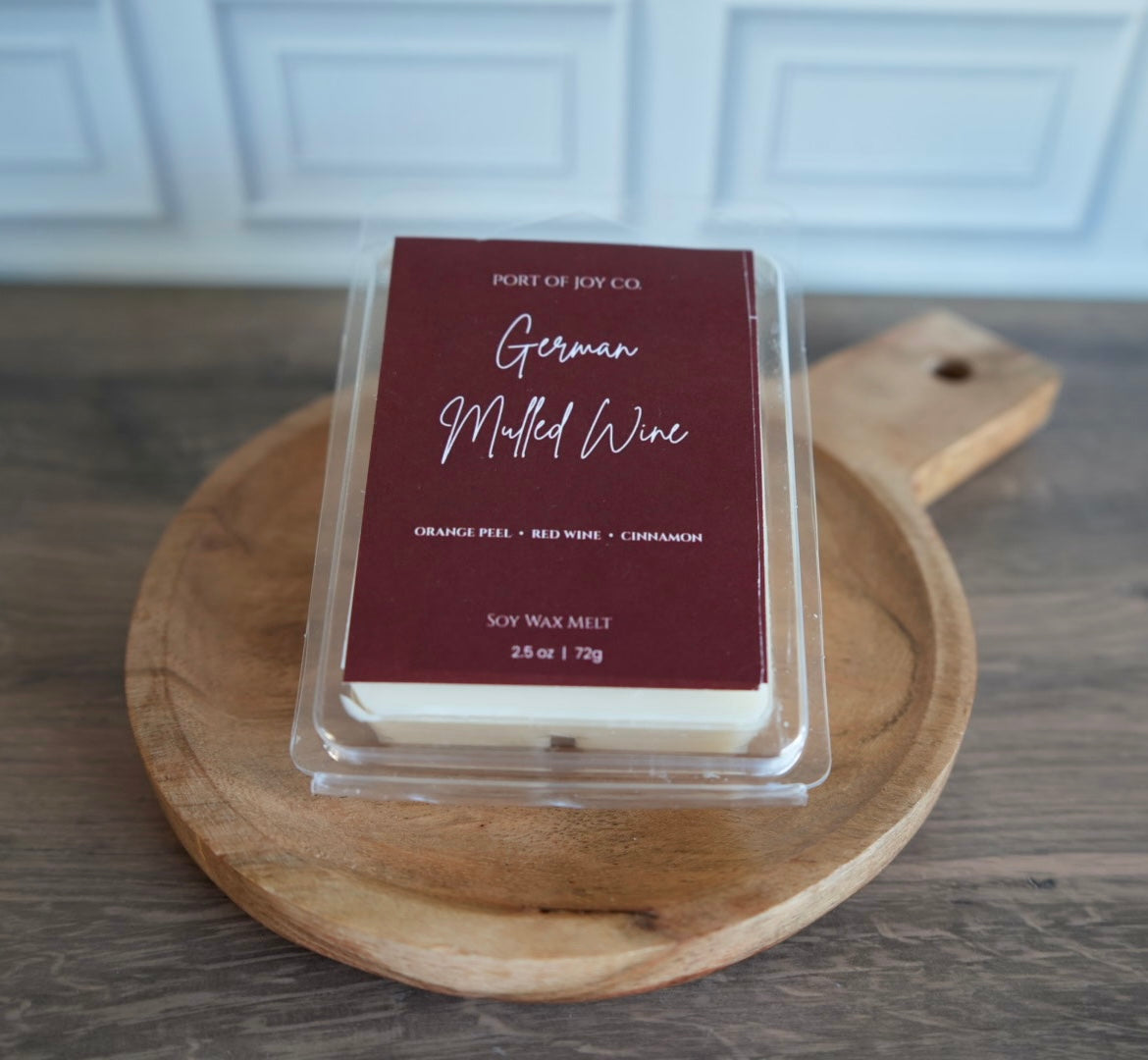 A clear clamshell package of Port of Joy Co. German Muddled Wine soy wax melt sits on a round wooden cutting board over a dark wood tabletop, with a blurred white paneled cabinet in the background.