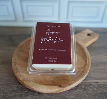 A clear clamshell package of Port of Joy Co. German Muddled Wine soy wax melt sits on a round wooden cutting board over a dark wood tabletop, with a blurred white paneled cabinet in the background.