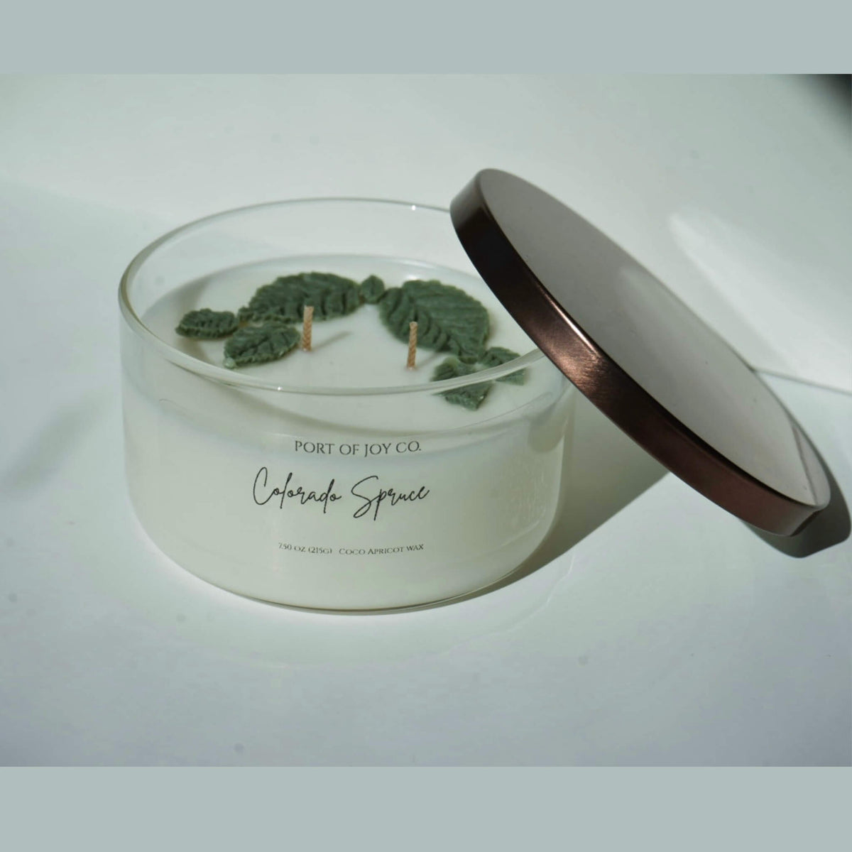 A clear glass jar candle with white wax and dried green leaves on top, shown with its lid leaning against it on a light surface. The label reads Port of Joy Co.