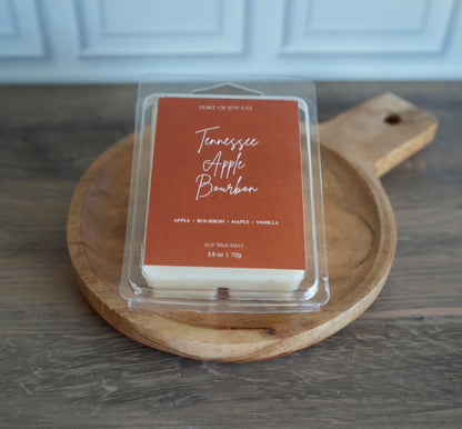 A packaged soy wax melt sits on a round wooden serving board on a gray wood tabletop.  apple, bourbon, maple, and vanilla.