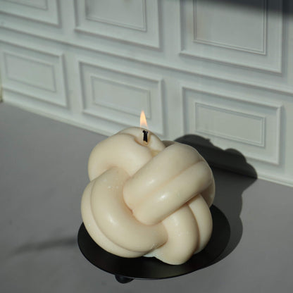 Knot-shaped candle on a black stand against a white paneled wall.
