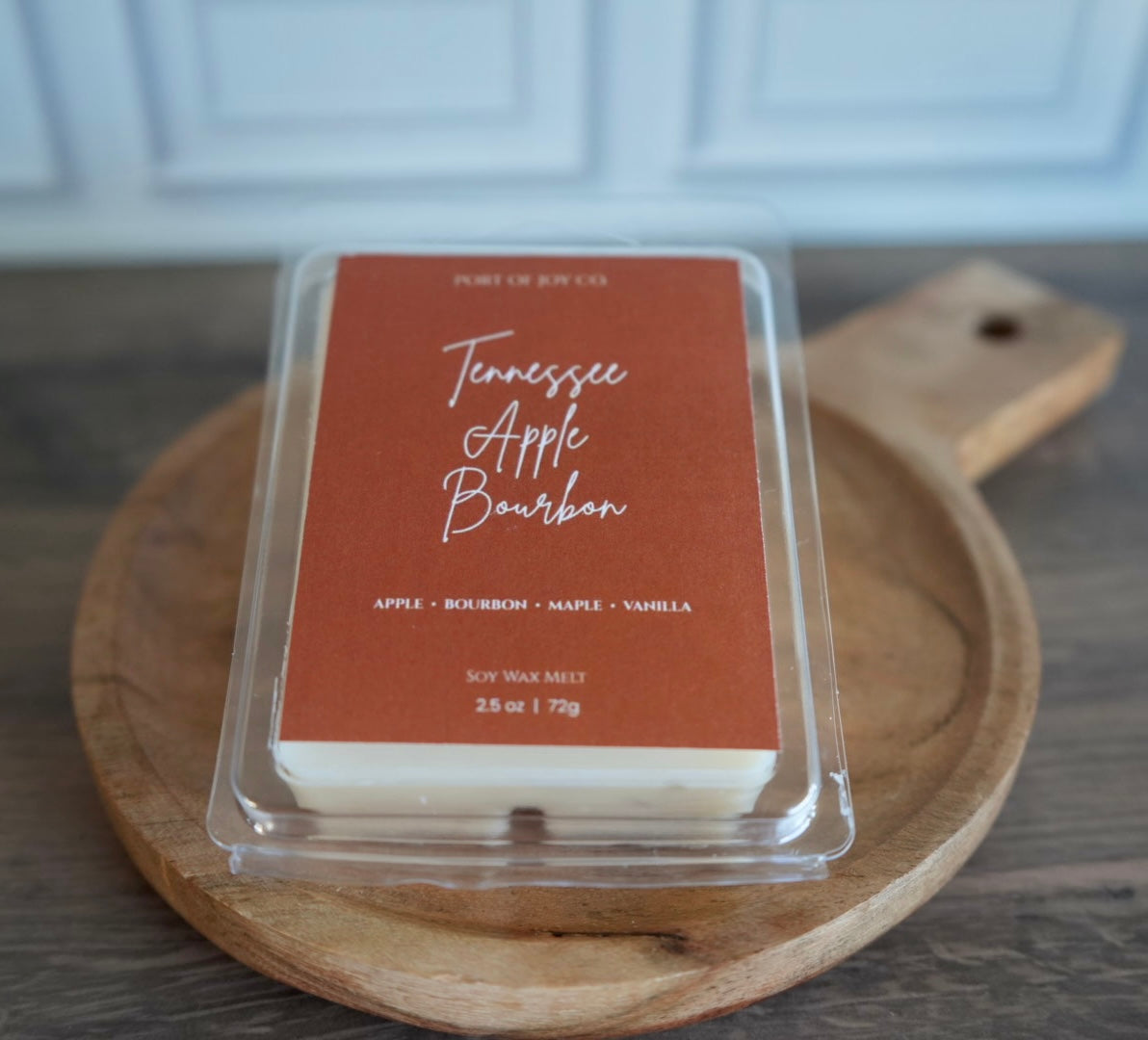 A clear clamshell package of Port of Joy Co. Tennessee Apple Bourbon soy wax melts sits on a round wooden serving board over a dark wood tabletop.