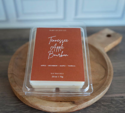 A clear clamshell package of Port of Joy Co. Tennessee Apple Bourbon soy wax melts sits on a round wooden serving board over a dark wood tabletop.