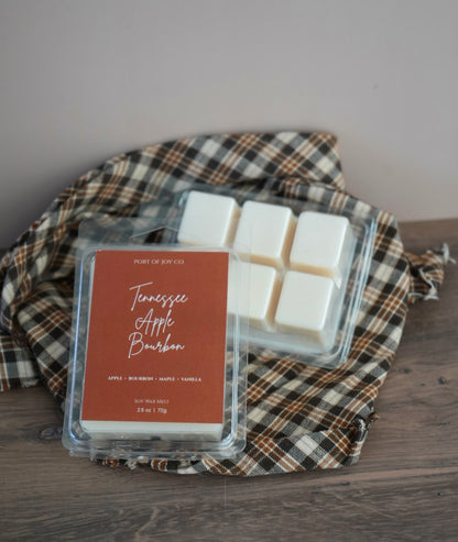 Two wax melts packages on a plaid cloth over a wooden surface. The front package has a label reading Port of Joy Co.