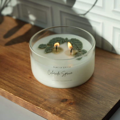 A double-wick scented candle in a clear glass jar sits lit on a wooden surface. 