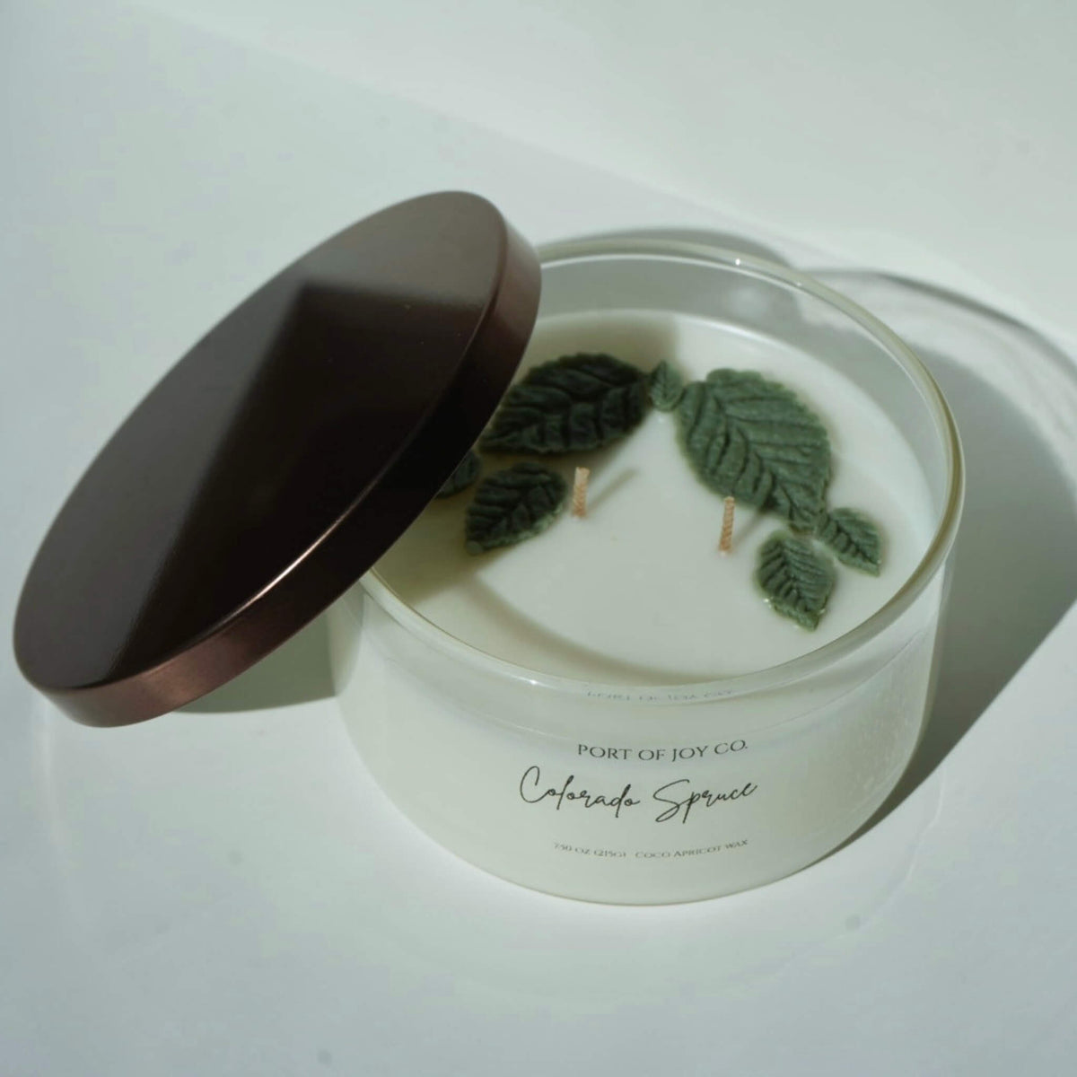 A white coconut wax candle in a clear glass jar with a dark brown lid leaning against it. Inside the jar are green leaf wicks and pressed green leaves.